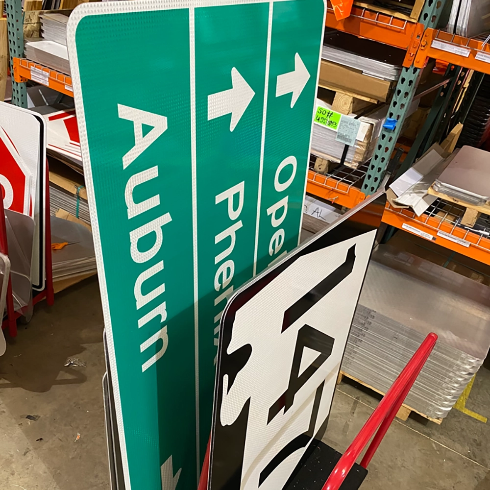 Large green retroreflective destination guide signs displaying white text and arrows for Auburn, Phenix City, and Opelika leaning against warehouse shelving at Rice Signs, with a black and white Alabama state route marker sign visible in the foreground.