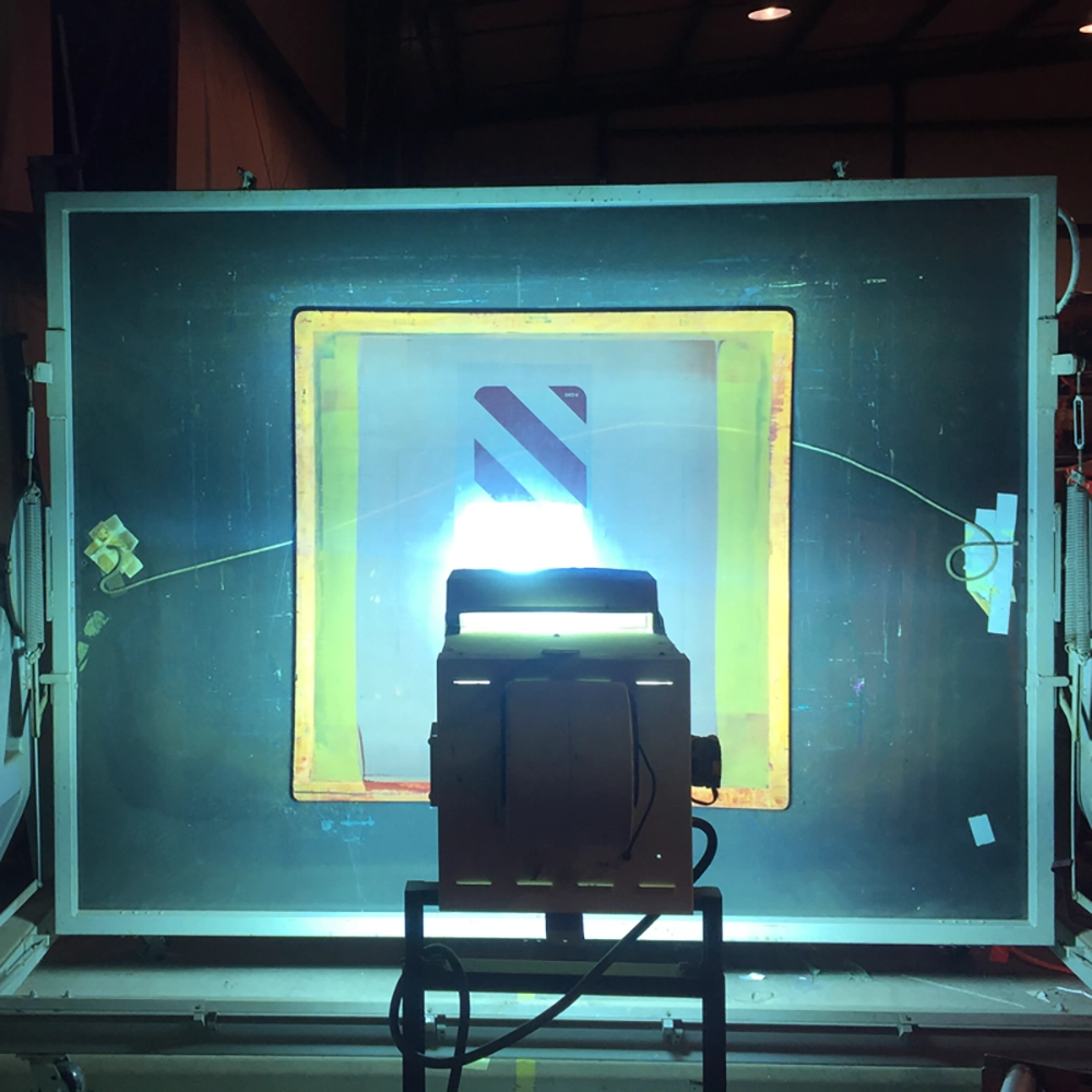 A large silk screen held flat inside a vacuum frame being exposed by an intense UV light source at the Rice Signs facility, with a diagonal stripe object marker pattern visible on the film positive centered on the screen mesh.