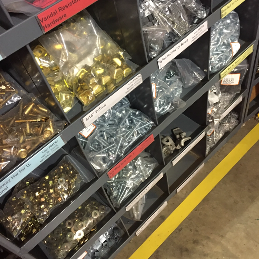 A gray metal parts bin organizer at Rice Signs filled with assorted sign mounting hardware including vandal-resistant bolts, 5/16-inch tufnuts, one-way bolts, nylon fence mounts, jam nuts, brass carriage bolts, and washers, each compartment labeled for quick identification.