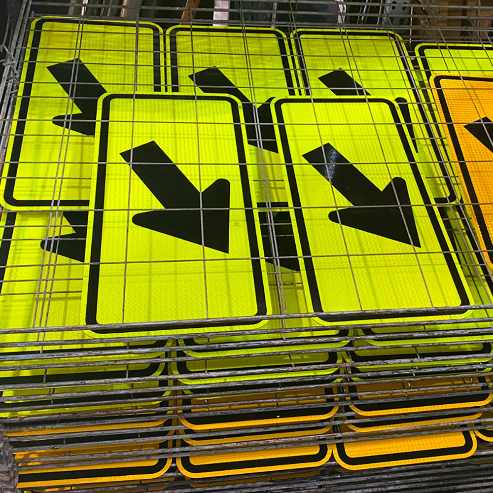 Stacks of fluorescent yellow-green W16-7P diagonal downward pointing arrow plaques drying on wire mesh racks after screen printing at the Rice Signs manufacturing facility, with black arrows and borders freshly applied to reflective sheeting.