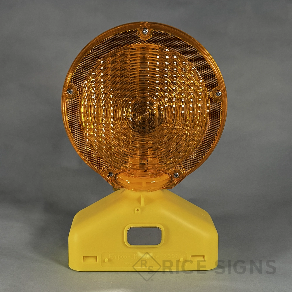 An Empco-Lite Model 2003 barricade warning light with a large round dual-sided amber Fresnel lens surrounded by an amber reflective border, secured with four visible screws, mounted on a yellow plastic base with a recessed handle slot and manufacturer labeling.