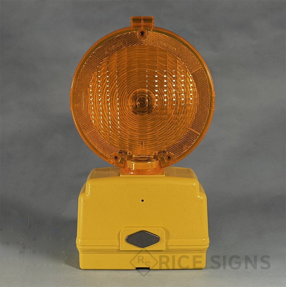 A battery-powered barricade warning light with a large round amber Fresnel lens and amber reflective border, mounted on a rectangular yellow plastic battery housing base featuring a diamond-shaped on/off switch on the front and a small photocell sensor hole, designed for mounting on traffic barricades and construction devices.