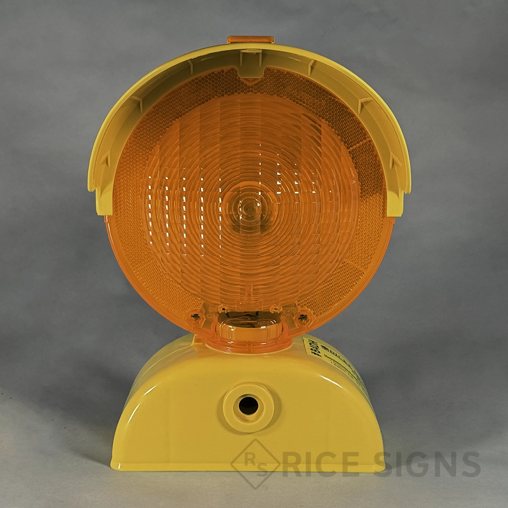 Front view of a battery-powered barricade warning light featuring a large round amber Fresnel lens with a concentric ring pattern, surrounded by an amber reflective border, housed in a yellow plastic shell with a hinged visor hood on top, a photocell sensor between the lens and base, and a yellow dome-shaped base with a central mounting hole for securing to barricades