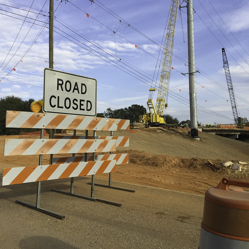 A road closure at an active bridge construction site, featuring a Type III barricade with three orange-and-white diagonally striped reflective rails, topped with a white 'ROAD CLOSED' sign and two amber warning lights. Behind the barricade, a large mound of red clay dirt leads to a bridge under construction, with a yellow crane and a dark lattice-boom crane operating on site. Power lines with red aerial marker balls span overhead, an orange traffic drum sits to the right, and the photo is taken from inside a vehicle with the driver's side mirror visible in the lower left corner.