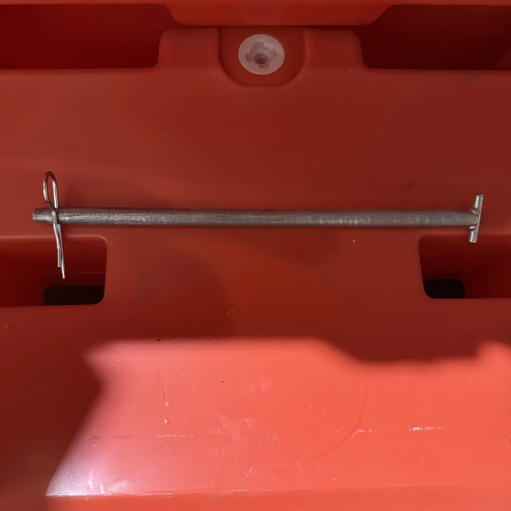 A steel connection pin with a cotter pin on one end and a cross pin on the other, stored horizontally in the built-in molded cradle on the side of an orange water-fill traffic barrier, with the white drain plug visible above.