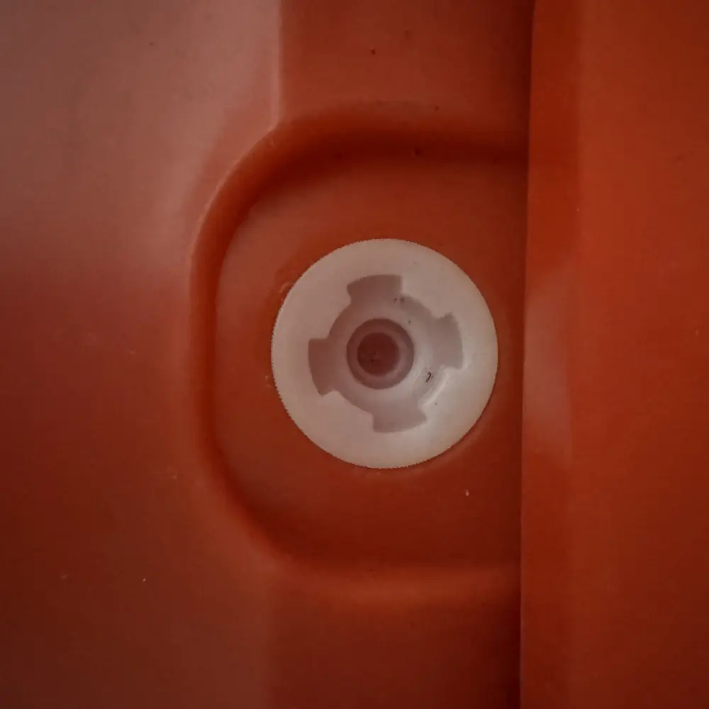 Close-up view of a white plastic drain plug seated in a recessed circular port on the side of an orange water-fill traffic barrier, featuring a cross-shaped grip pattern for hand-tightening to drain or seal the barrier.