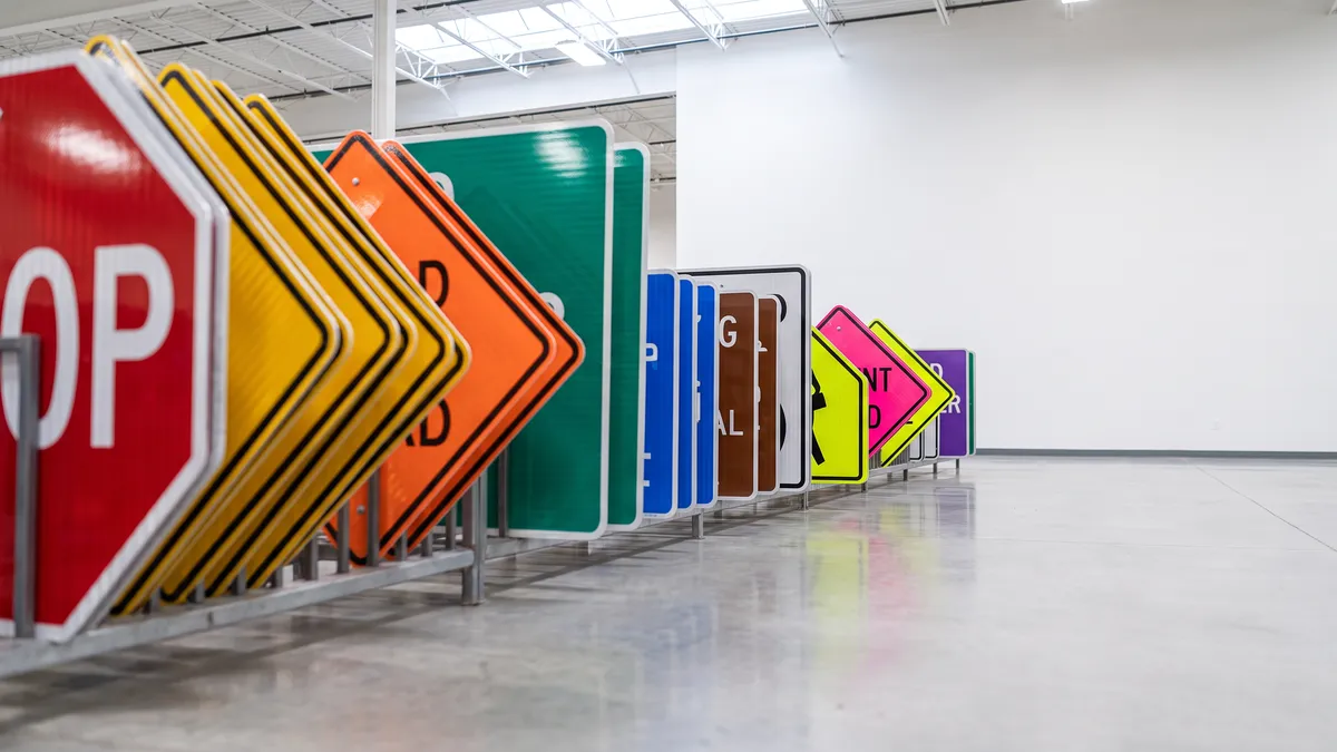Row of finished traffic signs in red, yellow, orange, green, blue, brown, white, fluorescent yellow-green, fluorescent pink and purple leaning on a rack in a bright clean sign warehouse.