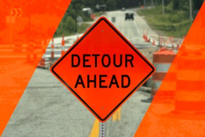 A large orange detour sign in a construction workzone with traffic drums and cones guiding traffic around the workers
